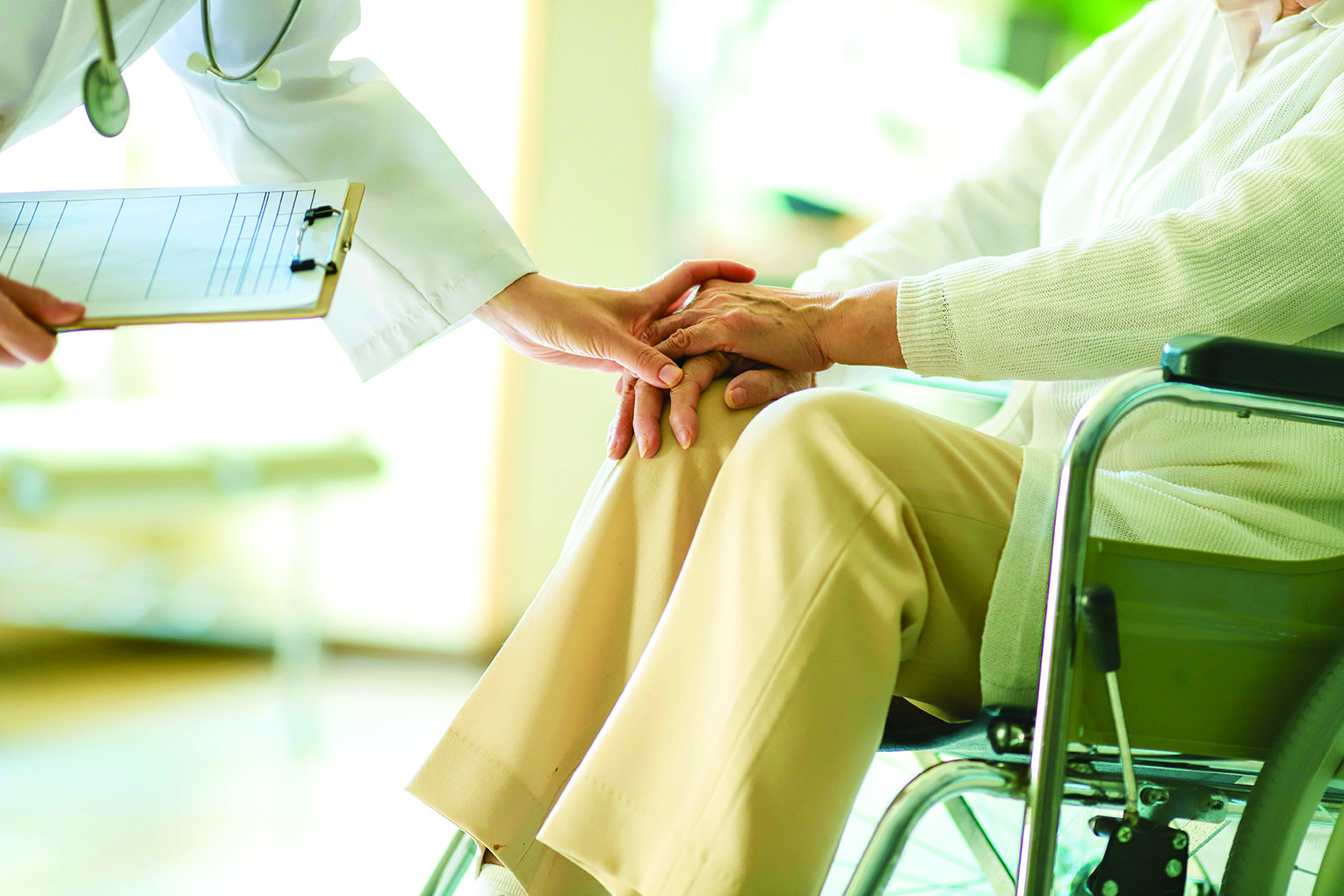 A clinician holds an elderly patient’s hand in a wheelchair, conveying care and support. A clipboard is visible nearby for notes.