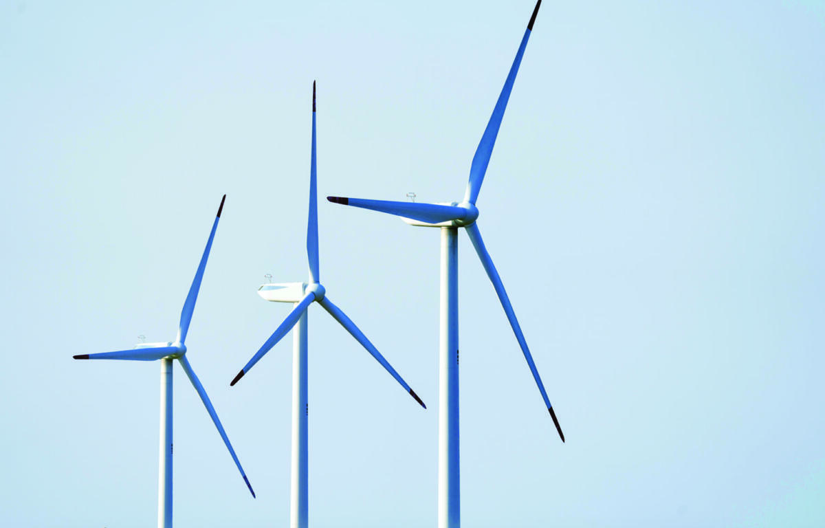 Three wind turbines with tall white towers and blue-blade rotors against a clear blue sky, illustrating renewable energy infrastructure.],
