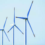Three wind turbines with tall white towers and blue-blade rotors against a clear blue sky, illustrating renewable energy infrastructure.],