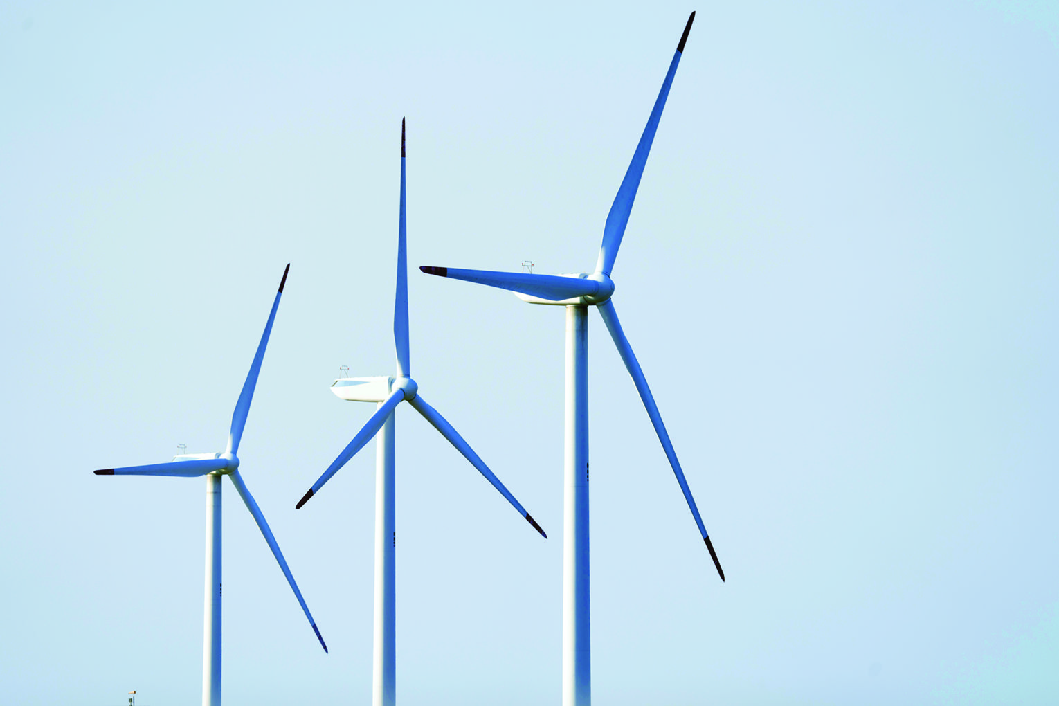 Three wind turbines with tall white towers and blue-blade rotors against a clear blue sky, illustrating renewable energy infrastructure.],