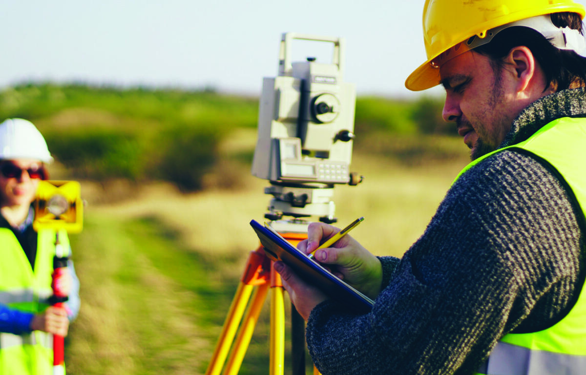 Two construction workers survey a site with a theodolite on a tripod while one takes notes on a clipboard.