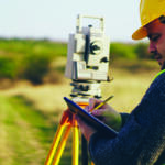 Two construction workers survey a site with a theodolite on a tripod while one takes notes on a clipboard.