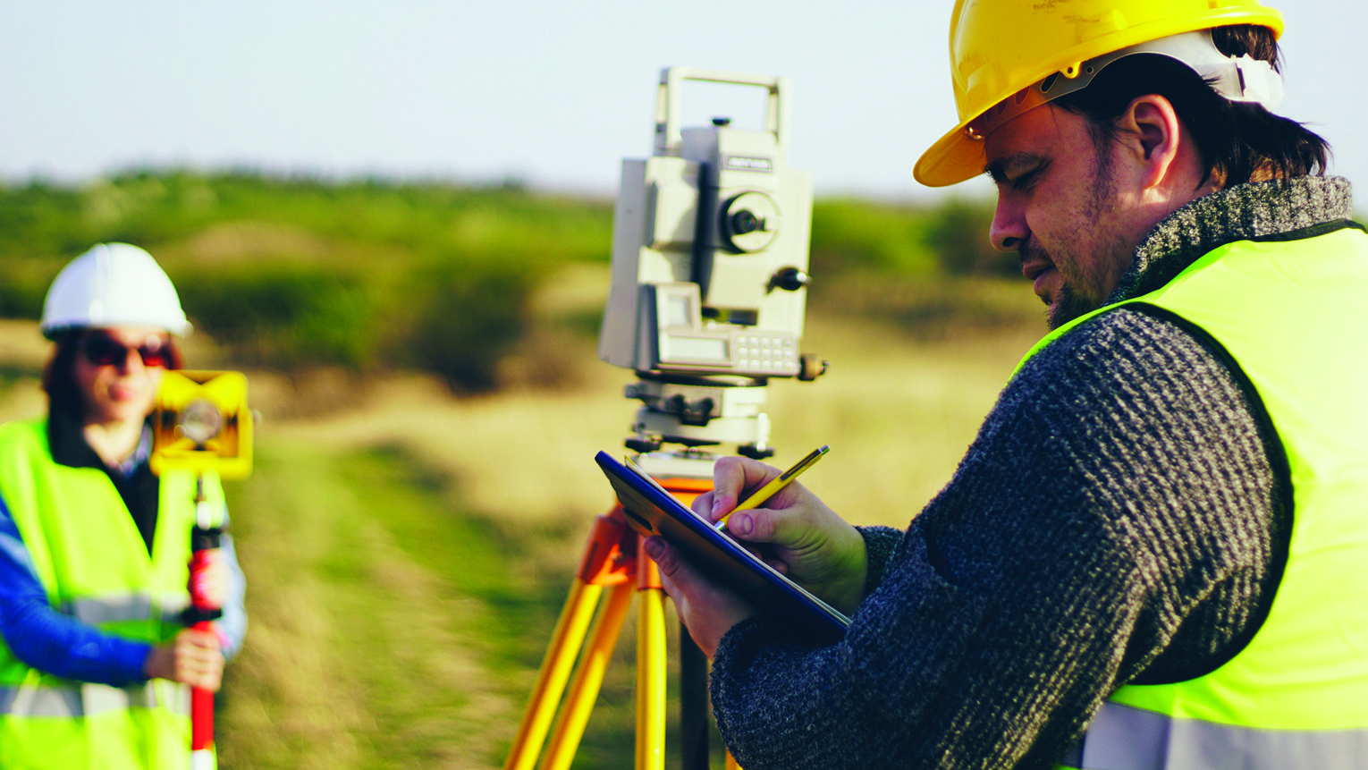Two construction workers survey a site with a theodolite on a tripod while one takes notes on a clipboard.