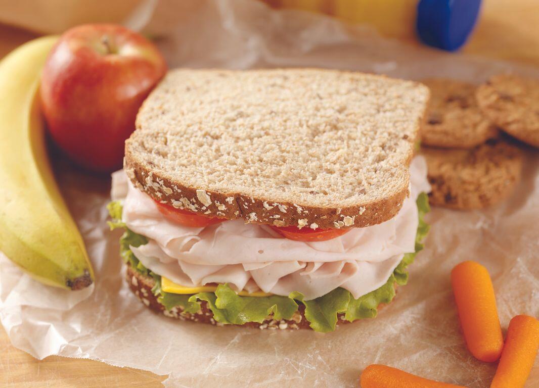 Whole‑grain turkey sandwich with lettuce and tomato on parchment, surrounded by fruit and carrot sticks.
