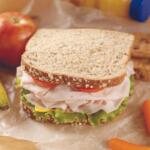 Whole‑grain turkey sandwich with lettuce and tomato on parchment, surrounded by fruit and carrot sticks.