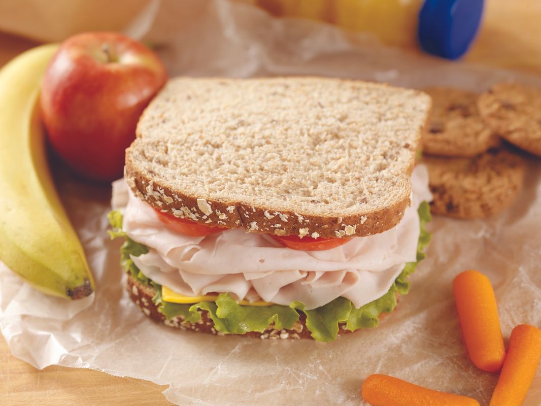 Whole‑grain turkey sandwich with lettuce and tomato on parchment, surrounded by fruit and carrot sticks.
