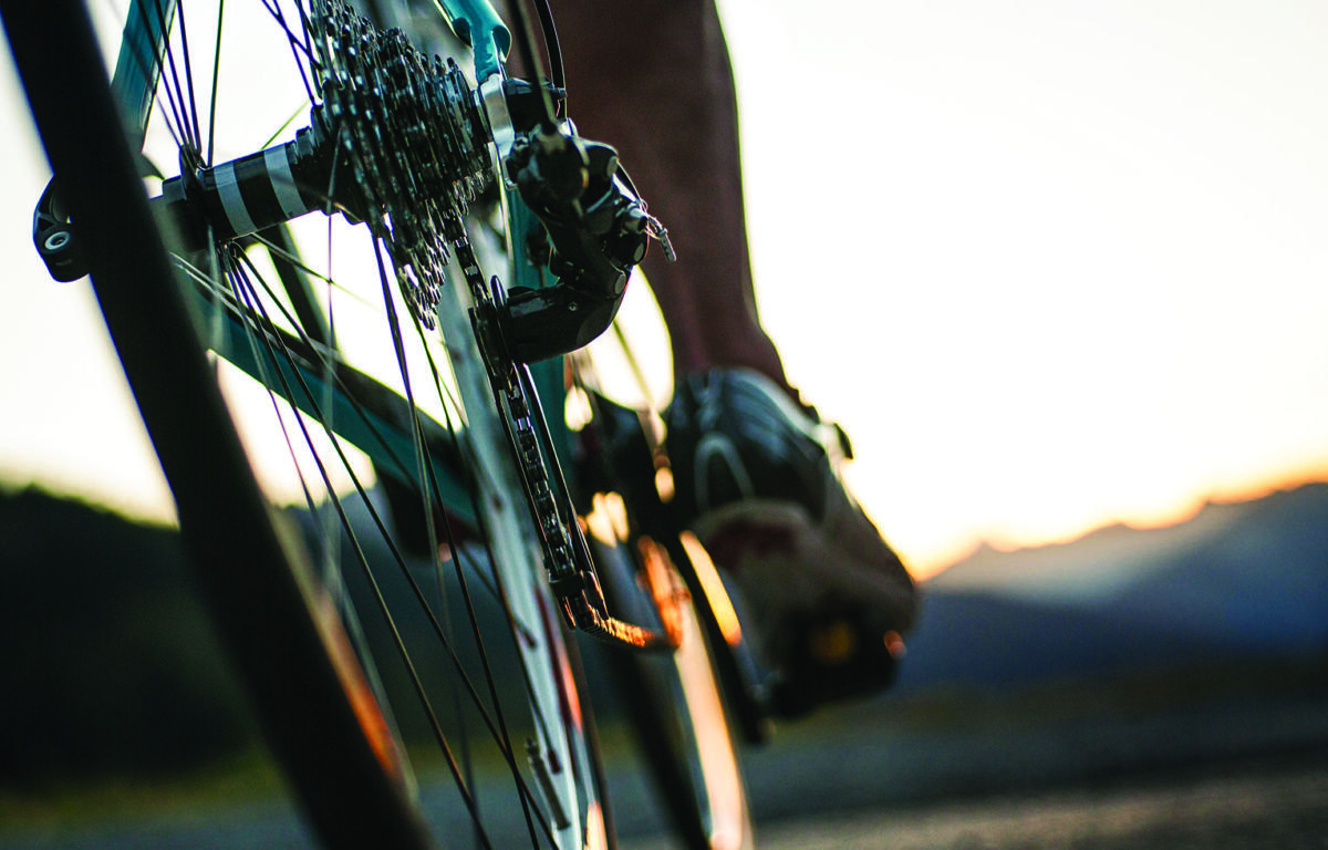 Close-up of a bicycle's rear derailleur and gears with a rider's foot on the pedal at sunset.