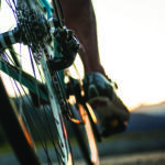 Close-up of a bicycle's rear derailleur and gears with a rider's foot on the pedal at sunset.