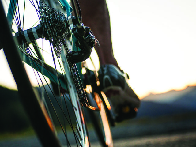 Close-up of a bicycle's rear derailleur and gears with a rider's foot on the pedal at sunset.