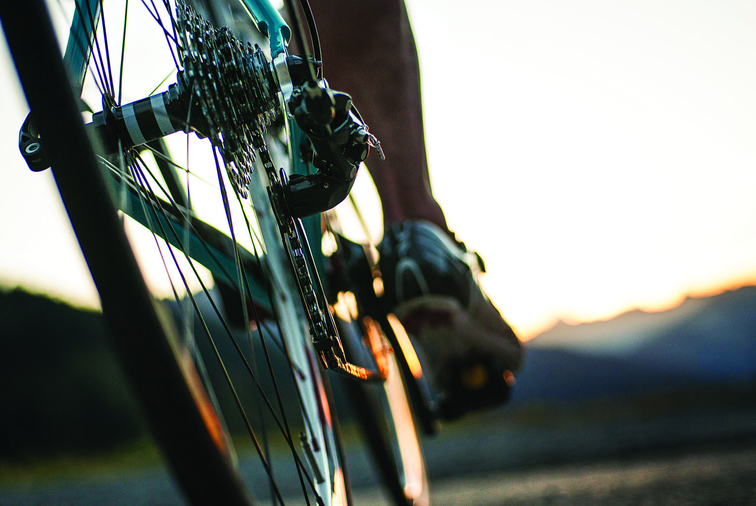 Close-up of a bicycle's rear derailleur and gears with a rider's foot on the pedal at sunset.