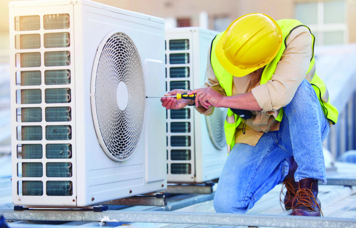 HVAC technician in a yellow hard hat and safety vest kneels on a rooftop, using a screwdriver on an outdoor air conditioning unit.
