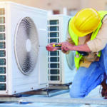 HVAC technician in a yellow hard hat and safety vest kneels on a rooftop, using a screwdriver on an outdoor air conditioning unit.