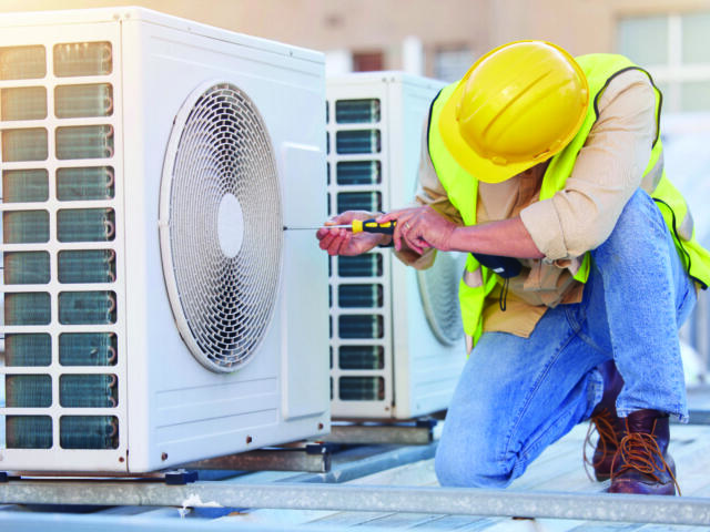 HVAC technician in a yellow hard hat and safety vest kneels on a rooftop, using a screwdriver on an outdoor air conditioning unit.