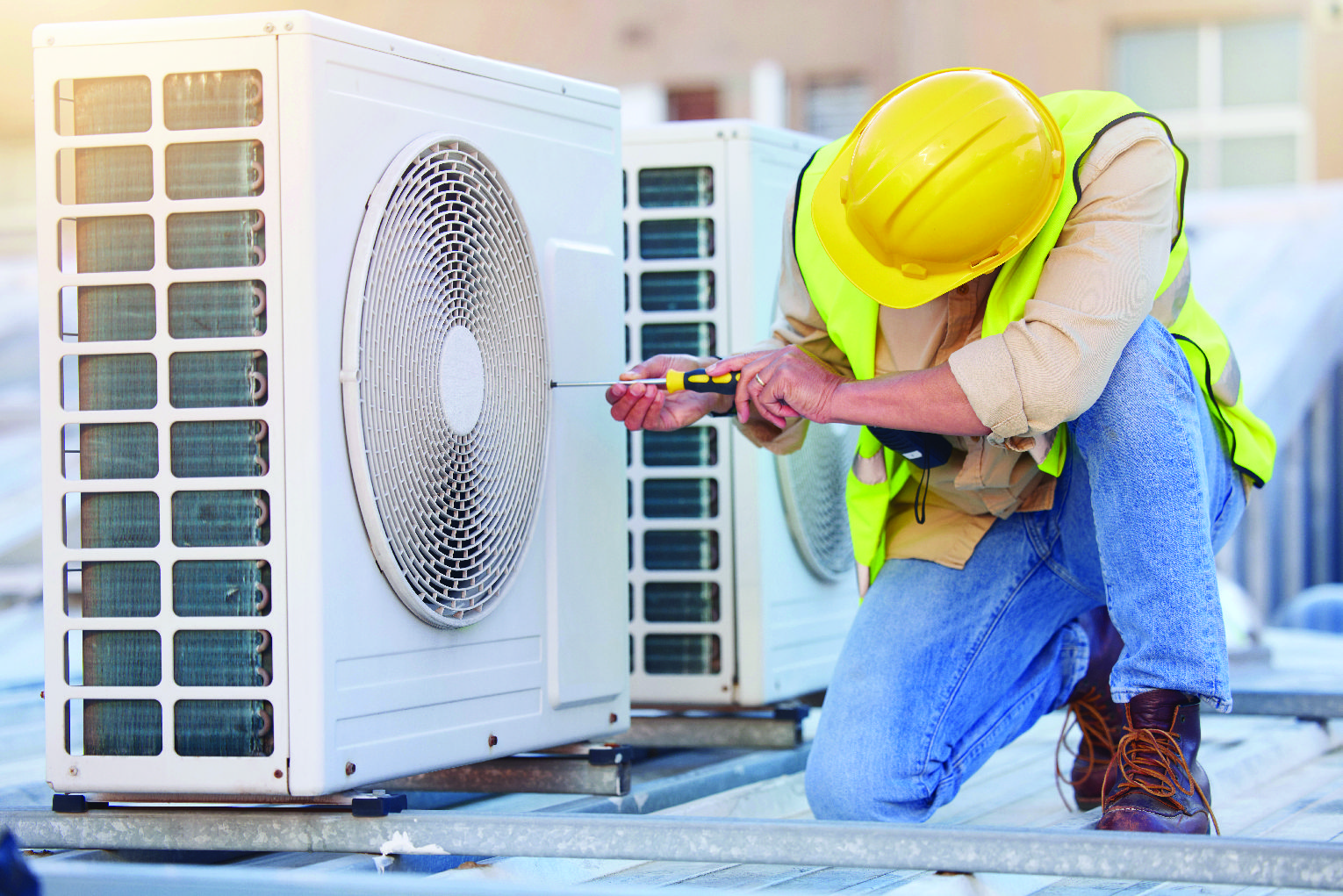 HVAC technician in a yellow hard hat and safety vest kneels on a rooftop, using a screwdriver on an outdoor air conditioning unit.
