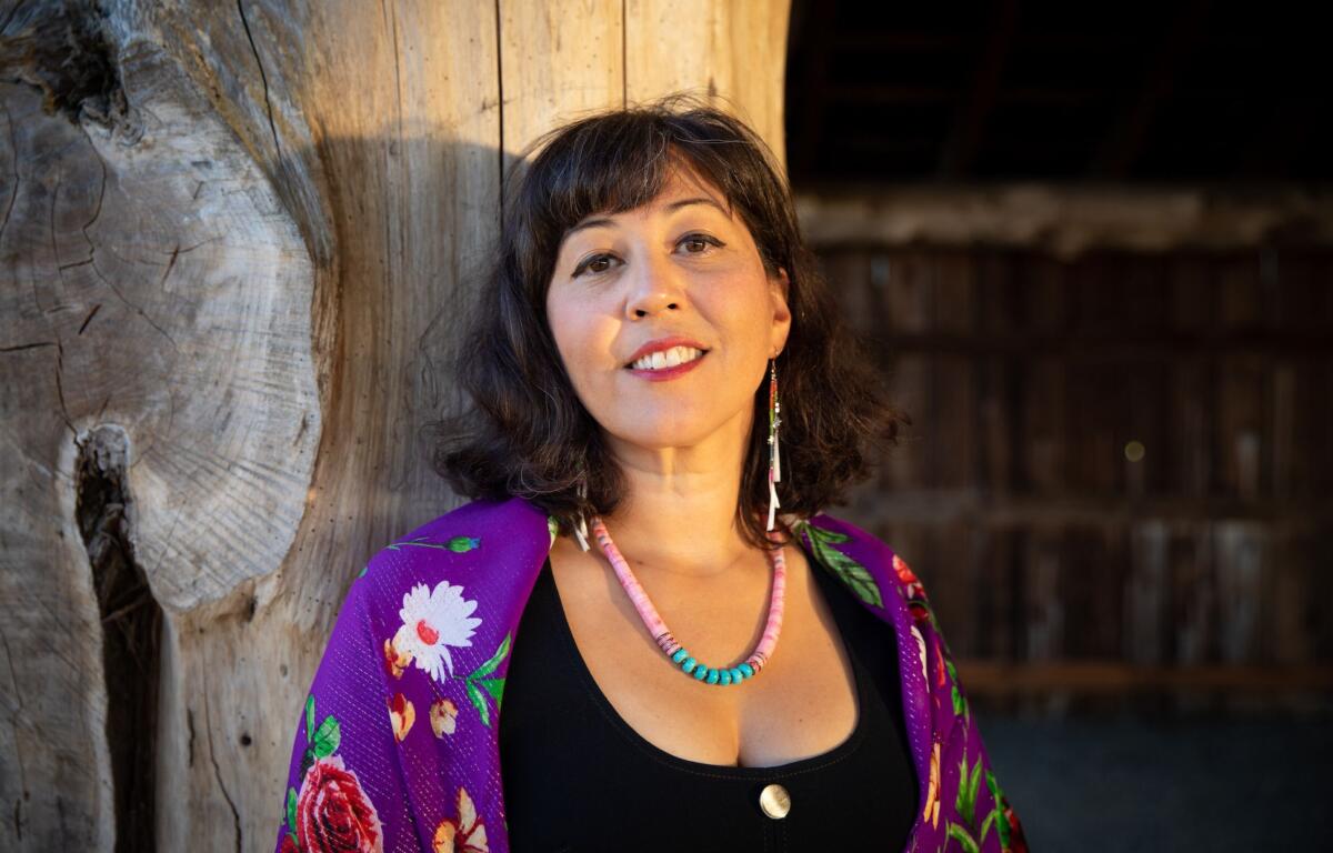 Woman leaning against a large weathered wooden post, smiling, wearing a purple floral shawl and turquoise bead necklace.