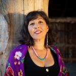Woman leaning against a large weathered wooden post, smiling, wearing a purple floral shawl and turquoise bead necklace.