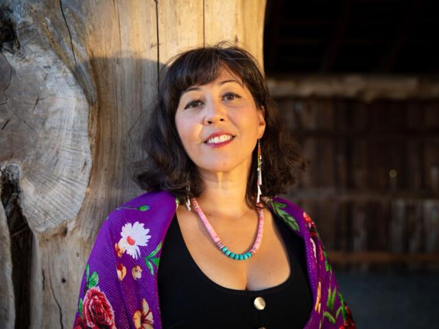 Woman leaning against a large weathered wooden post, smiling, wearing a purple floral shawl and turquoise bead necklace.