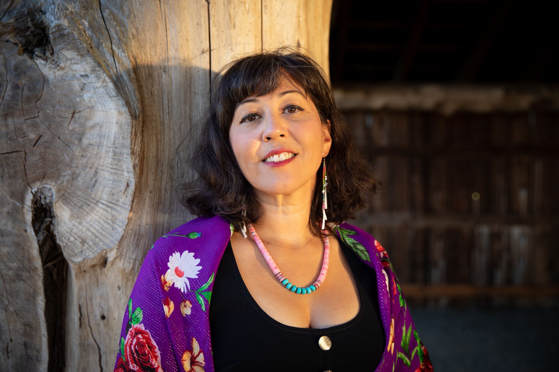 Woman leaning against a large weathered wooden post, smiling, wearing a purple floral shawl and turquoise bead necklace.