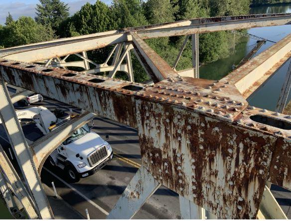 Close-up of rusted steel bridge girders with rivets, overlooking a road with a white truck beneath and trees in the background