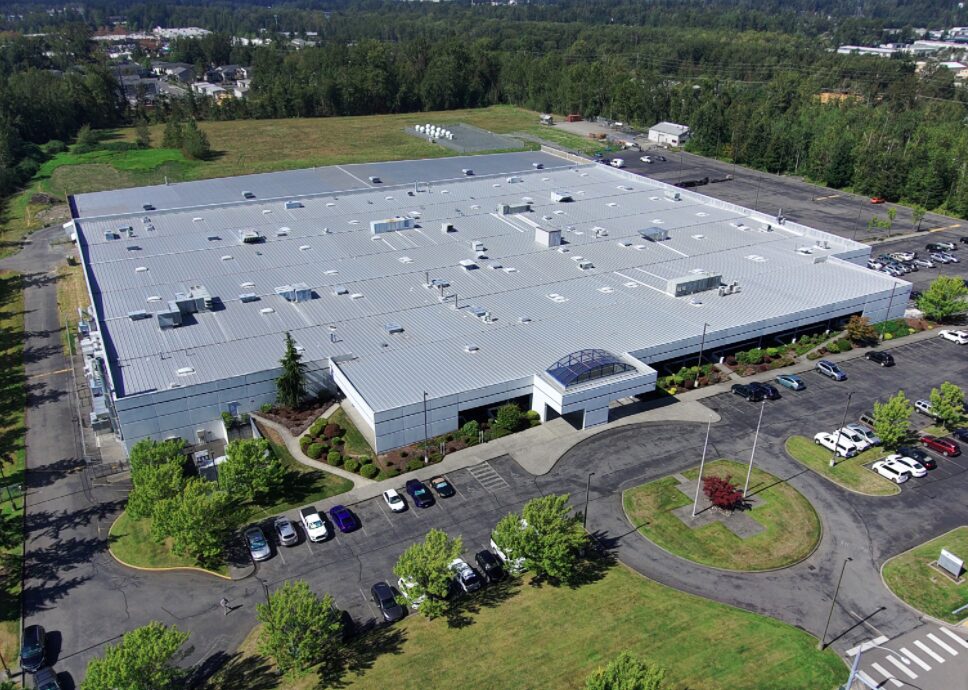 Aerial view of a large gray-roofed office/industrial building with a circular driveway and surrounding parking lot.
