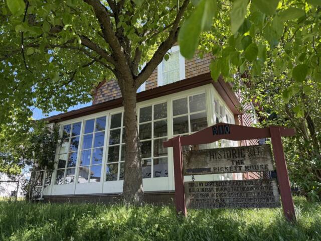 Two-story house with large white-framed windows, shaded by a leafy tree; a weathered sign reads 'Historic Site, The Pickett House'.