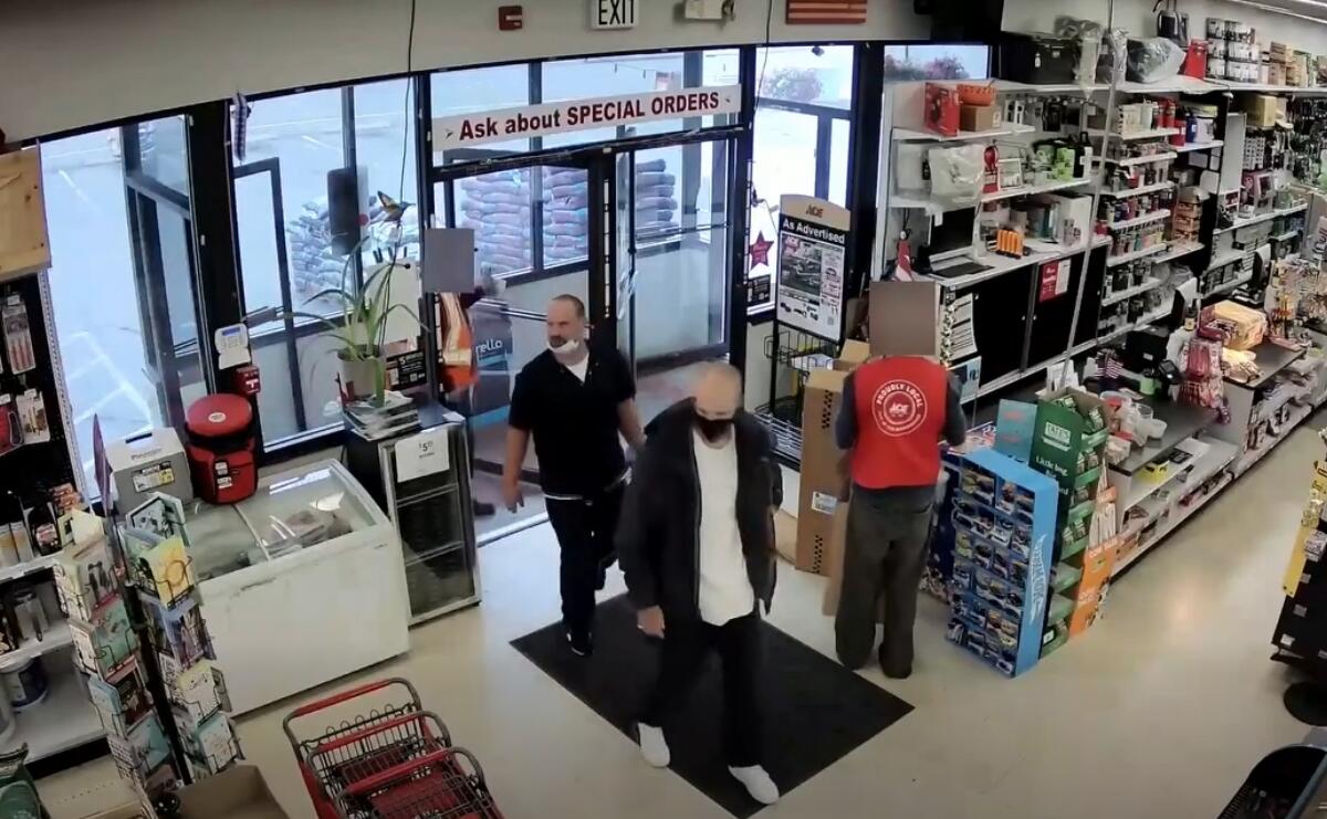 Two customers wearing masks stand near the entrance of a store while a staff member in a red vest works behind the counter near tall shelves.