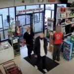 Two customers wearing masks stand near the entrance of a store while a staff member in a red vest works behind the counter near tall shelves.