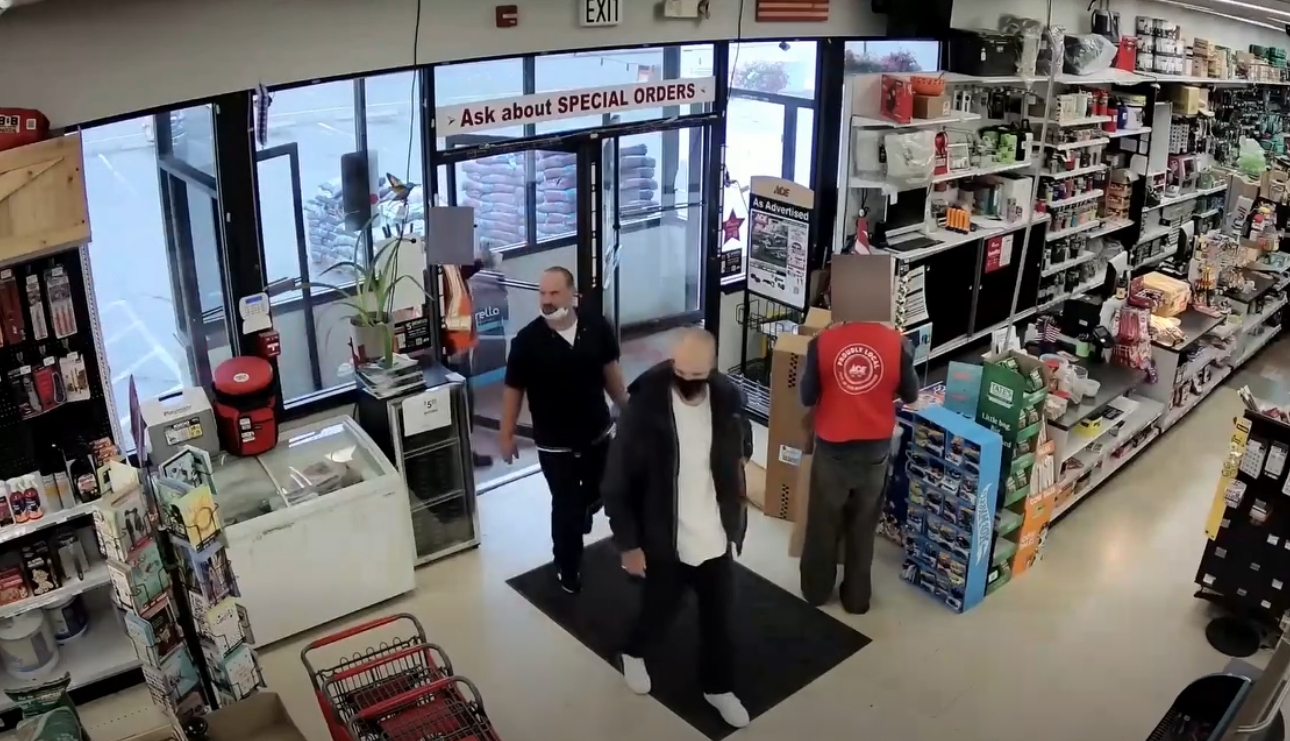 Two customers wearing masks stand near the entrance of a store while a staff member in a red vest works behind the counter near tall shelves.