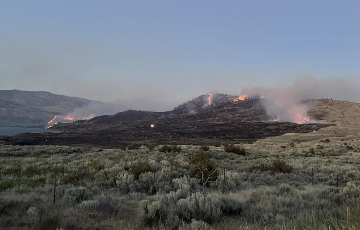 Hillsides burning with orange flames and smoke rising over a dry grassland near a lake, with a fence in the foreground.