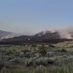 Hillsides burning with orange flames and smoke rising over a dry grassland near a lake, with a fence in the foreground.