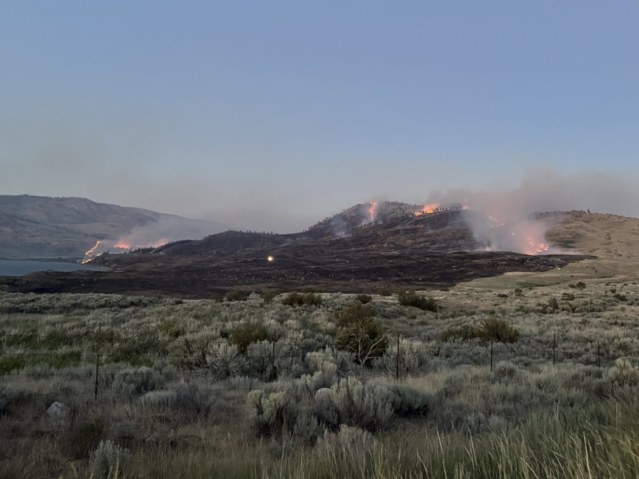 Hillsides burning with orange flames and smoke rising over a dry grassland near a lake, with a fence in the foreground.