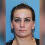 Mugshot-style photo of a woman with dark hair tied back, neutral expression, against a blue background; hoop earrings visible.