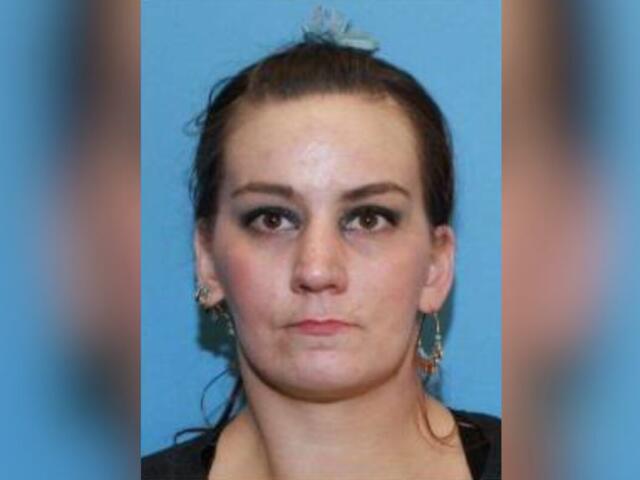 Mugshot-style photo of a woman with dark hair tied back, neutral expression, against a blue background; hoop earrings visible.