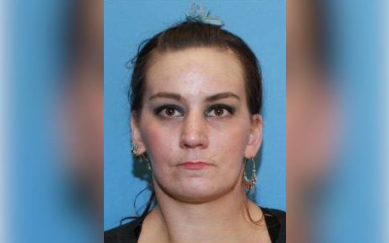 Mugshot-style photo of a woman with dark hair tied back, neutral expression, against a blue background; hoop earrings visible.