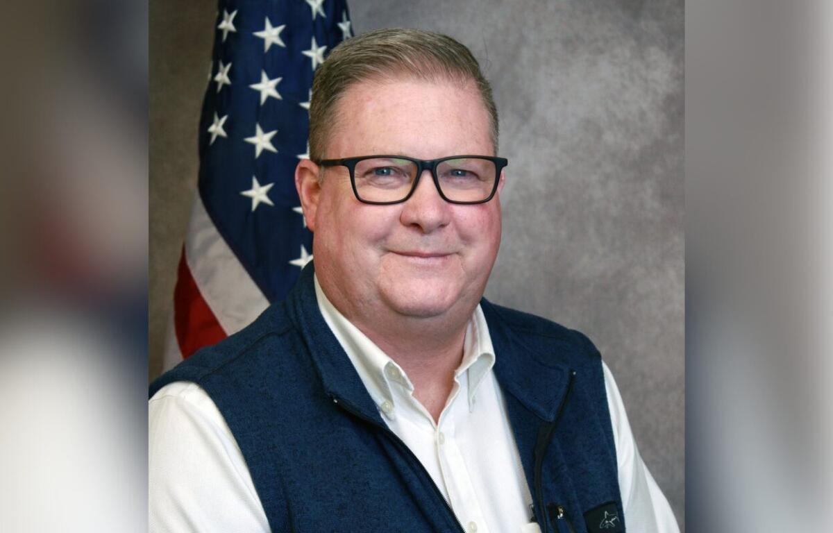 Portrait of a smiling man wearing glasses, a white shirt and navy vest, with an American flag in the background.