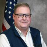 Portrait of a smiling man wearing glasses, a white shirt and navy vest, with an American flag in the background.
