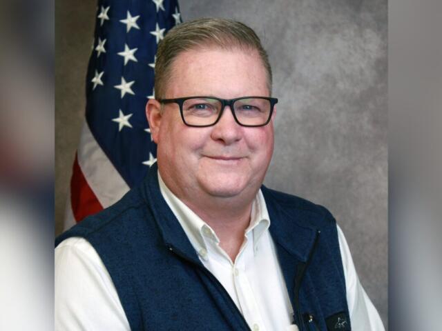 Portrait of a smiling man wearing glasses, a white shirt and navy vest, with an American flag in the background.