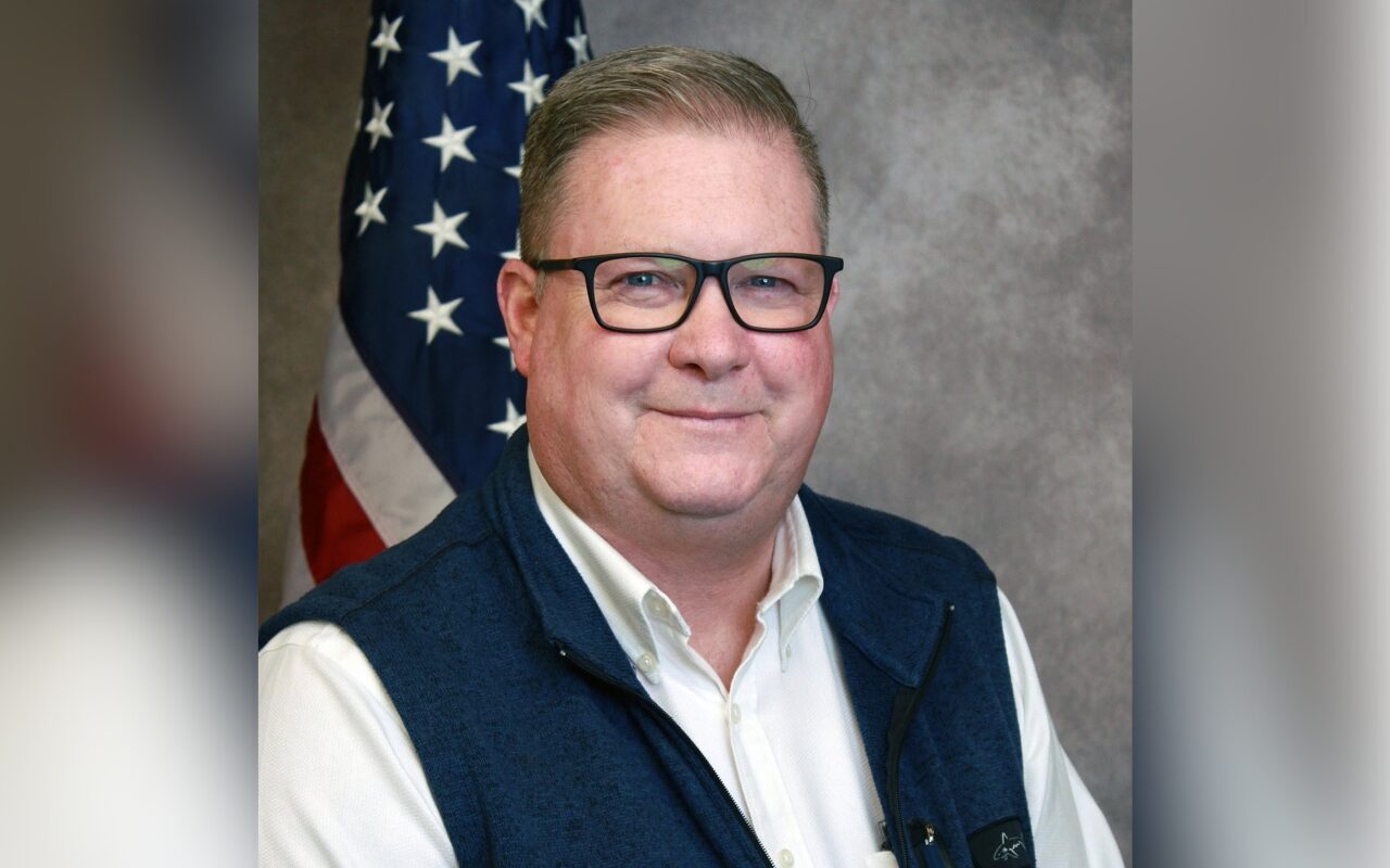 Portrait of a smiling man wearing glasses, a white shirt and navy vest, with an American flag in the background.