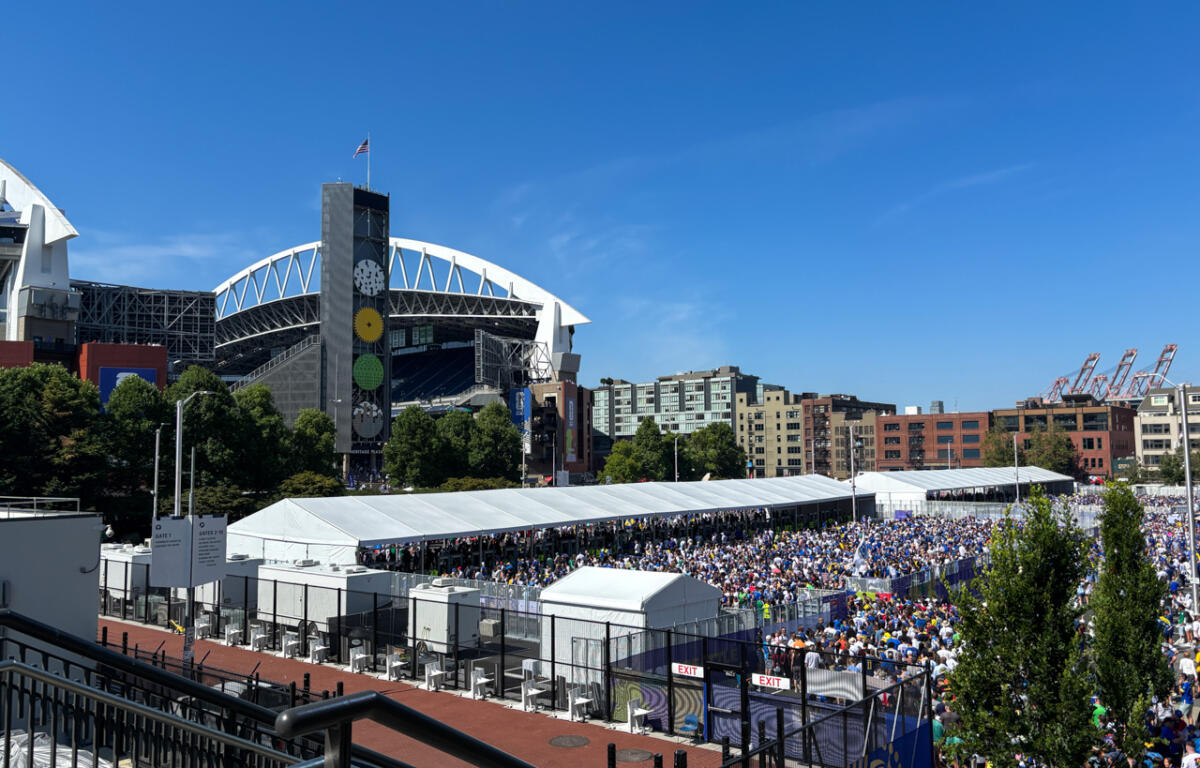 Crowd-filled outdoor event under long white tents beside a large modern stadium on a bright day, with a blue sky above.