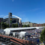 Crowd-filled outdoor event under long white tents beside a large modern stadium on a bright day, with a blue sky above.