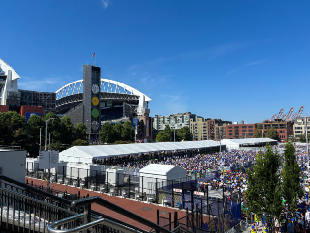 Crowd-filled outdoor event under long white tents beside a large modern stadium on a bright day, with a blue sky above.