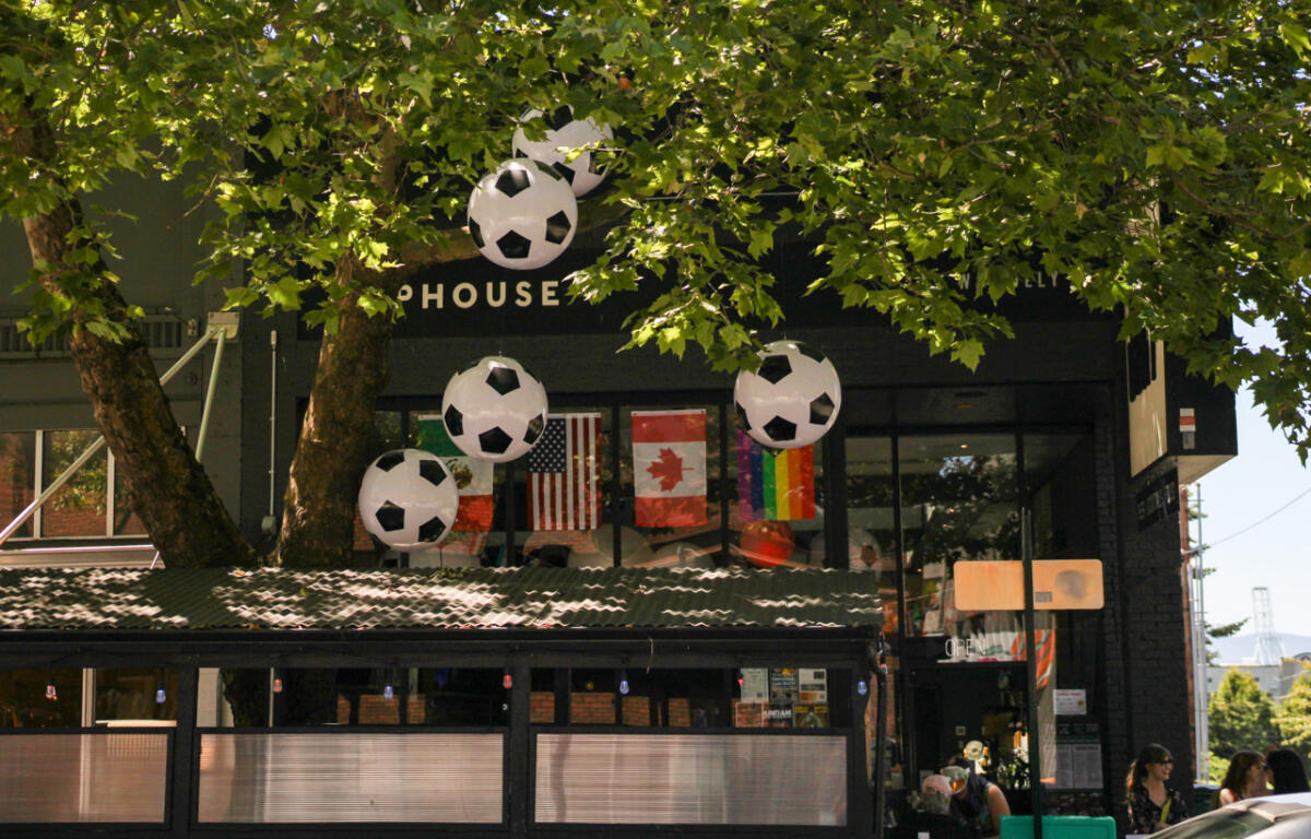 Storefront adorned with large hanging soccer-ball balloons, flags in the window, and people seated outside under leafy trees.