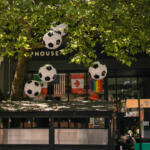 Storefront adorned with large hanging soccer-ball balloons, flags in the window, and people seated outside under leafy trees.