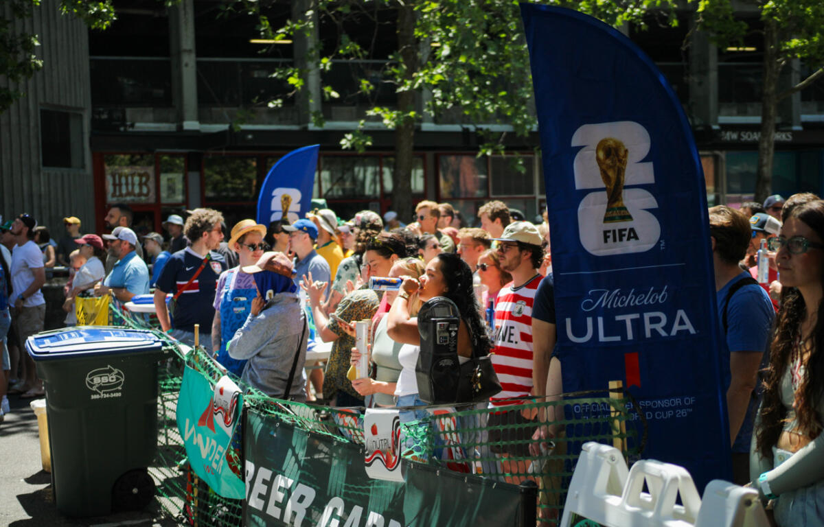 Crowd of spectators behind a green barricade at a sunny outdoor event, with a tall blue FIFA Michelob Ultra banner nearby.