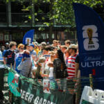 Crowd of spectators behind a green barricade at a sunny outdoor event, with a tall blue FIFA Michelob Ultra banner nearby.