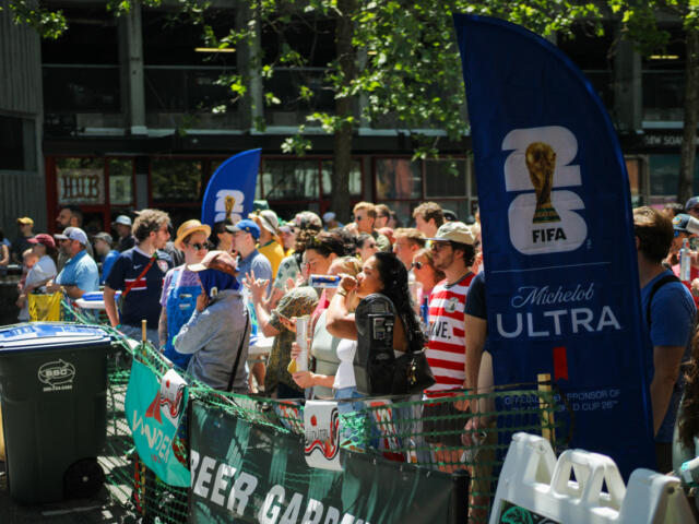 Crowd of spectators behind a green barricade at a sunny outdoor event, with a tall blue FIFA Michelob Ultra banner nearby.