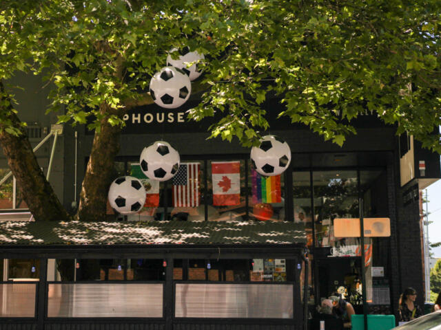 Storefront adorned with large hanging soccer-ball balloons, flags in the window, and people seated outside under leafy trees.