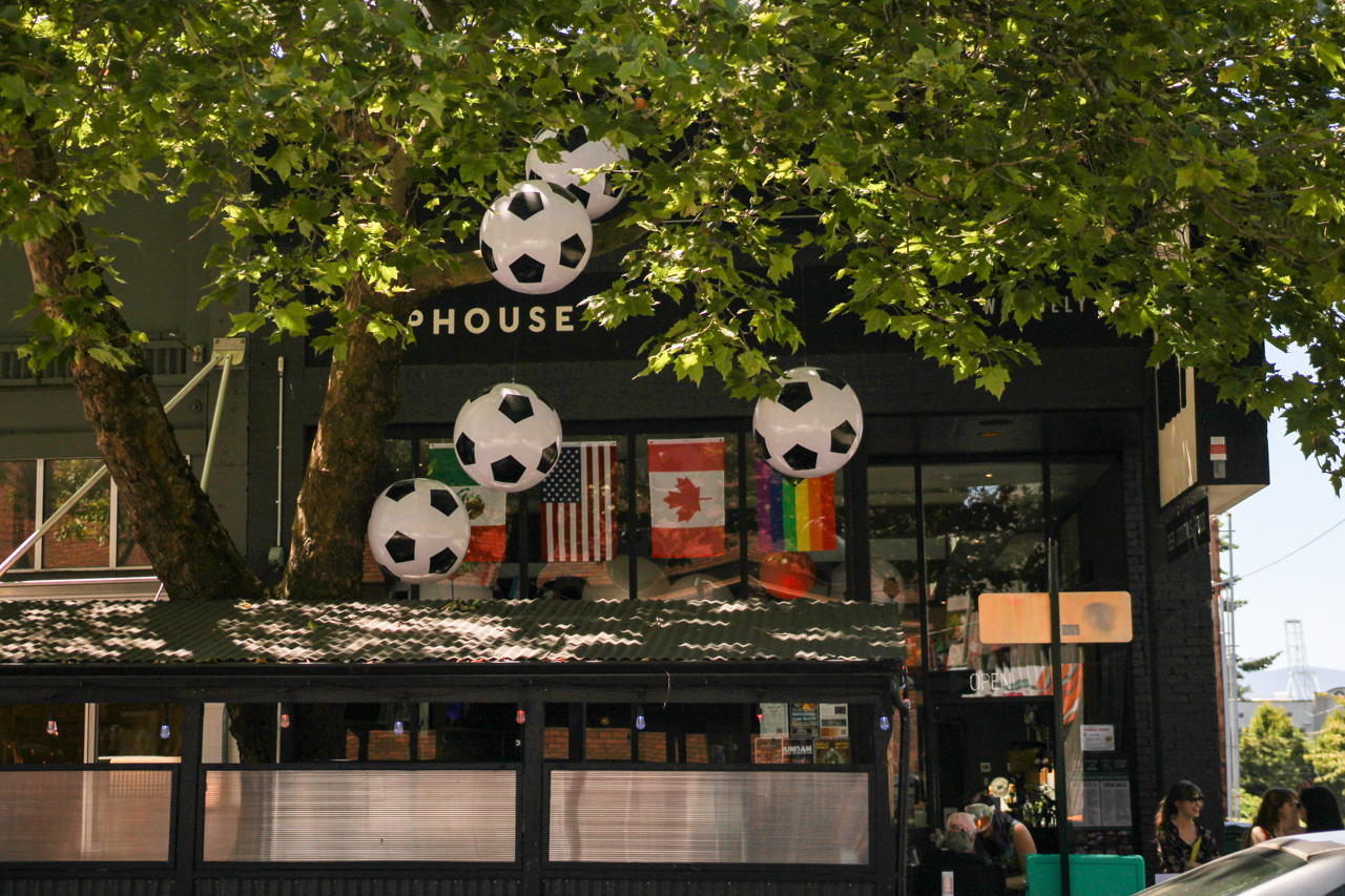 Storefront adorned with large hanging soccer-ball balloons, flags in the window, and people seated outside under leafy trees.