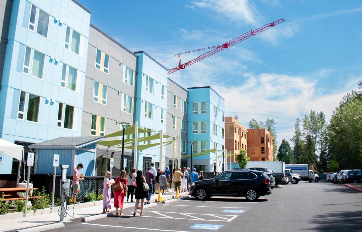 Group of people walking and standing in a parking lot beside a blue-gray modern building under a red construction crane in the sky.