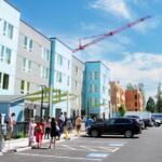 Group of people walking and standing in a parking lot beside a blue-gray modern building under a red construction crane in the sky.